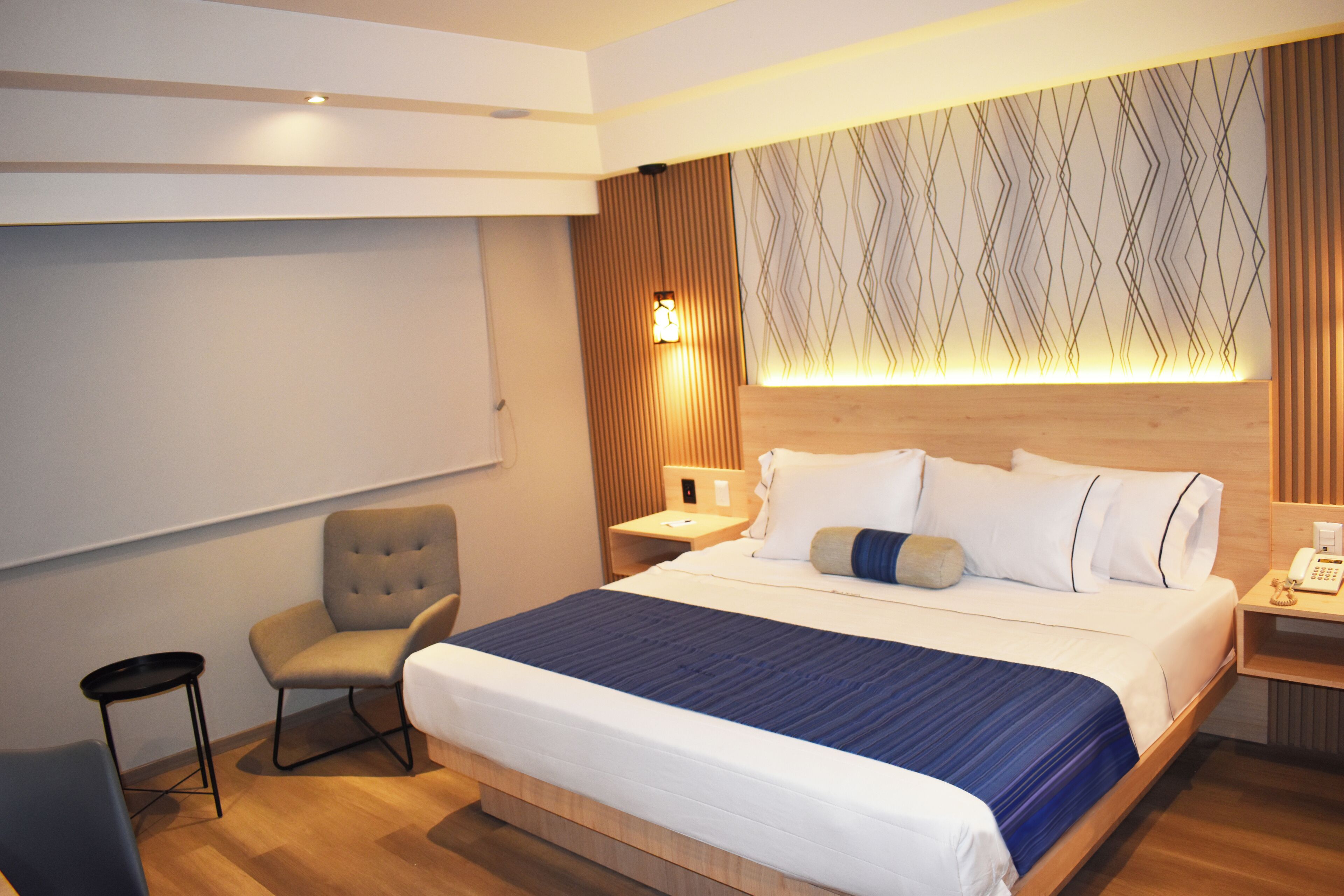 A hotel room with a large bed, a chair, a small table, and a nightstand with a lamp.