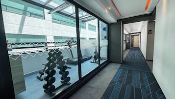 A corridor with a gym visible through a glass wall, a patterned carpet, and a hallway leading to a door.