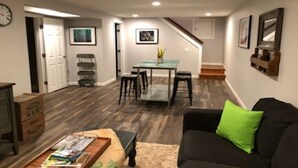 Una sala de estar con un sofá negro, una mesa de centro con revistas, un comedor con mesa de vidrio y una escalera que conduce a otro nivel.