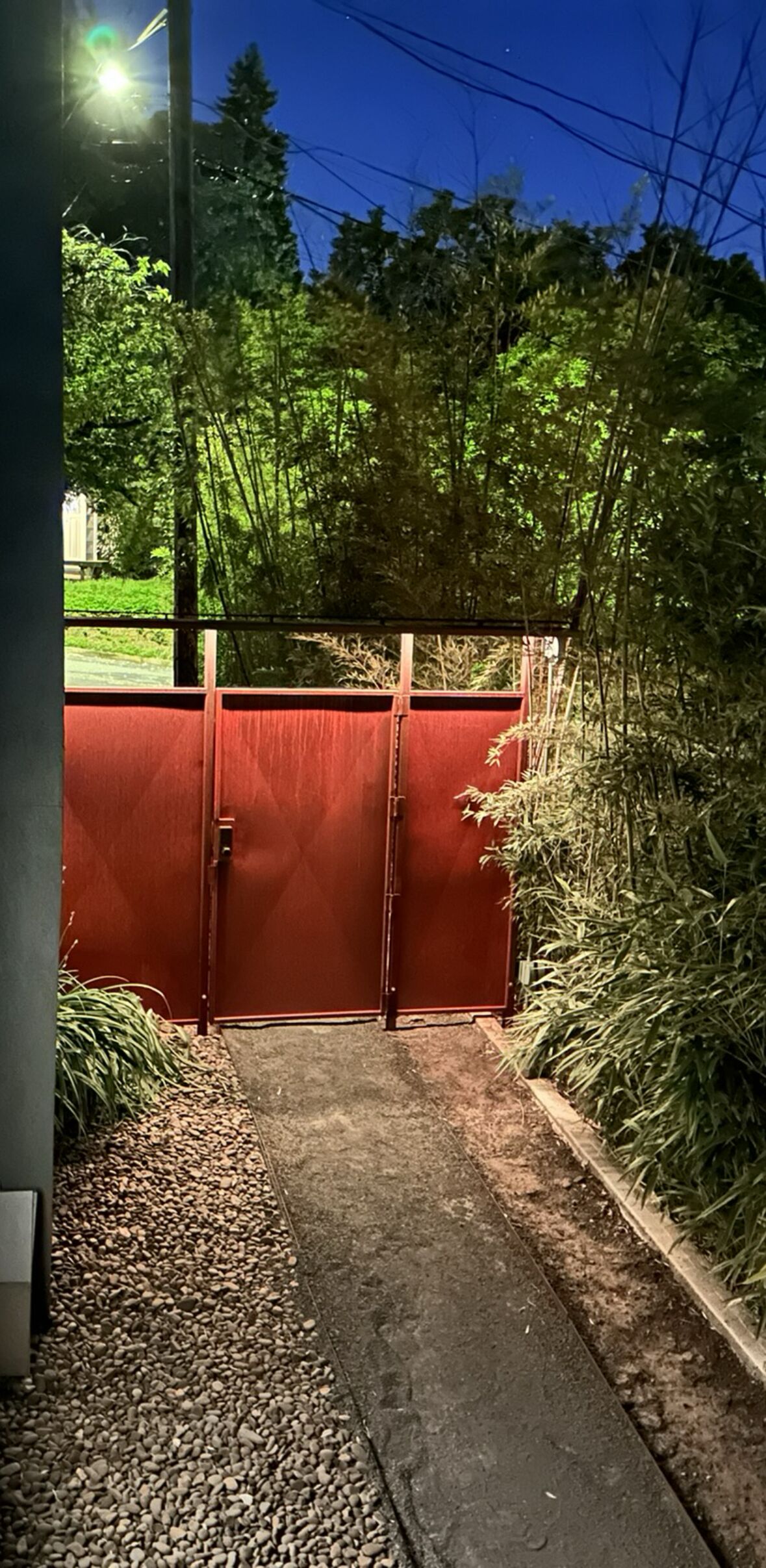 Una entrada protegida por un portón rojo, rodeada de follaje denso y un camino de grava.