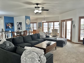 A spacious living room with a dark gray sectional sofa, a wooden coffee table, and a ceiling fan. There is a dining area with wooden chairs and a table, and large windows providing natural light.