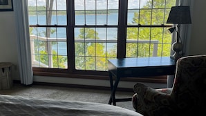 A room with a large window offering a view of a lake and trees. There is a bed, a chair, and a lamp on a table. - Luxury Waterfront Resort Home with Private Beach! (Rogers)