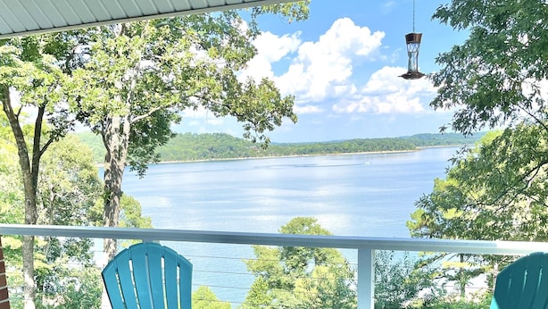A balcony with a glass table and two blue Adirondack chairs, overlooking a lake with trees and a clear sky. - Luxury Waterfront Resort Home with Private Beach! (Rogers)