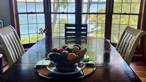 A dining table with a bowl of fruit in the center, surrounded by chairs, with a view of a body of water through large windows. - Luxury Waterfront Resort Home with Private Beach! (Rogers)
