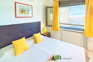 A hotel room with a bed, two yellow pillows, a framed artwork on the wall, a window with yellow curtains, and a view of the sea and boats.
