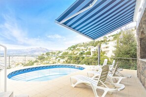 A swimming pool with lounge chairs, a blue-striped awning, and a view of a hillside town.