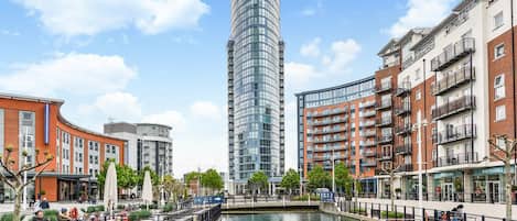 Un quartier urbain moderne, caractérisé par un bâtiment aux façades courbes en verre, un canal, ainsi que des immeubles résidentiels et commerciaux environnants.