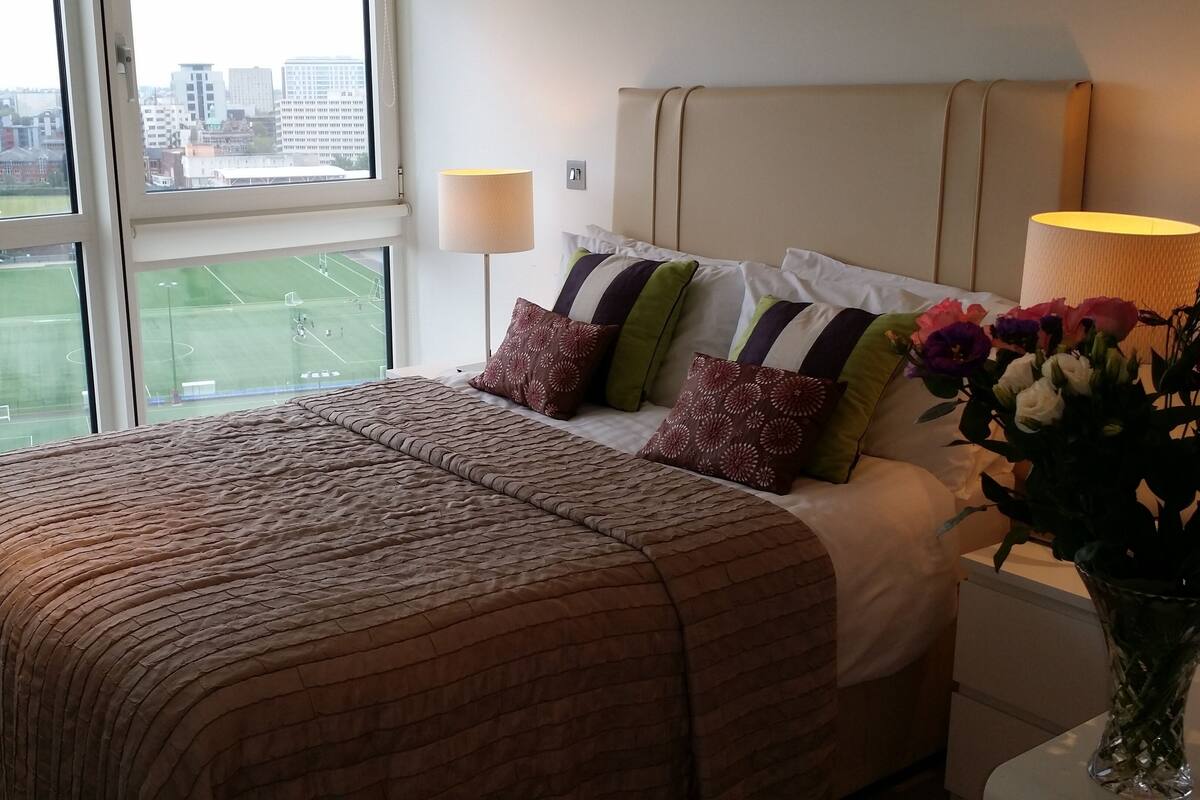 Une chambre d’hôtel avec un lit, une table de chevet, une lampe, un vase avec des fleurs et une fenêtre donnant sur un terrain de sport et des immeubles.