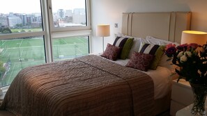 Ein Hotelzimmer mit Bett, Nachttisch, Lampe, einer Vase mit Blumen und einem Fenster mit Blick auf ein Sportfeld und Gebäude.