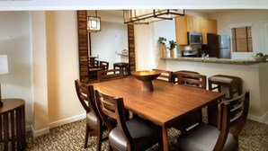 Un espace repas avec une table et des chaises en bois, un grand saladier au centre, et une cuisine en arrière-plan.