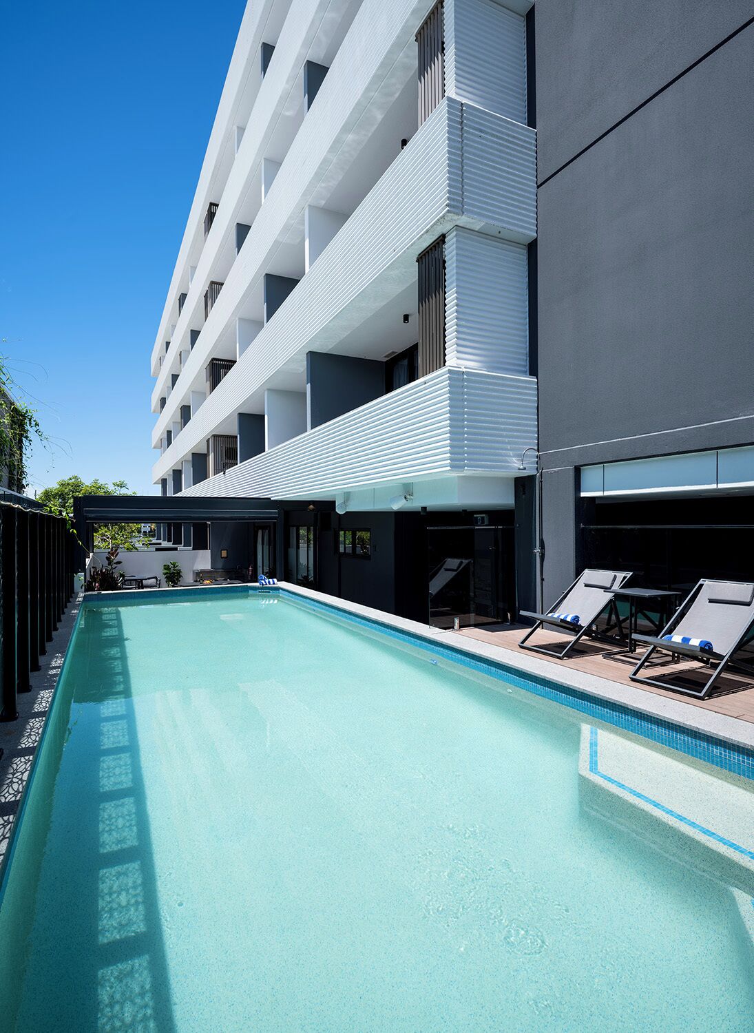 Une piscine avec des chaises longues, un bâtiment doté de balcons et un ciel dégagé.