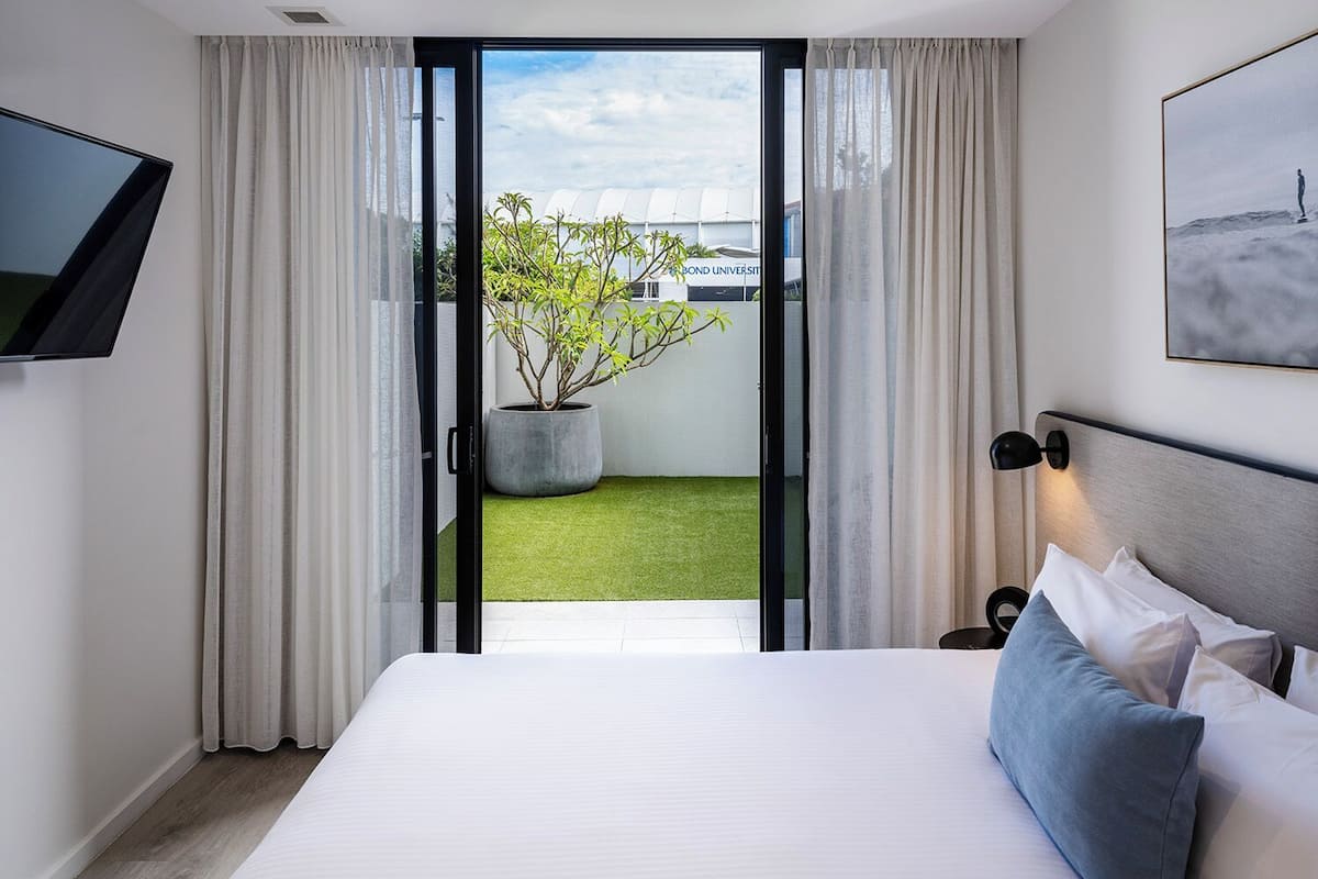 Habitación de hotel con cama, televisor montado en la pared, planta en maceta y vistas a un edificio y vegetación.