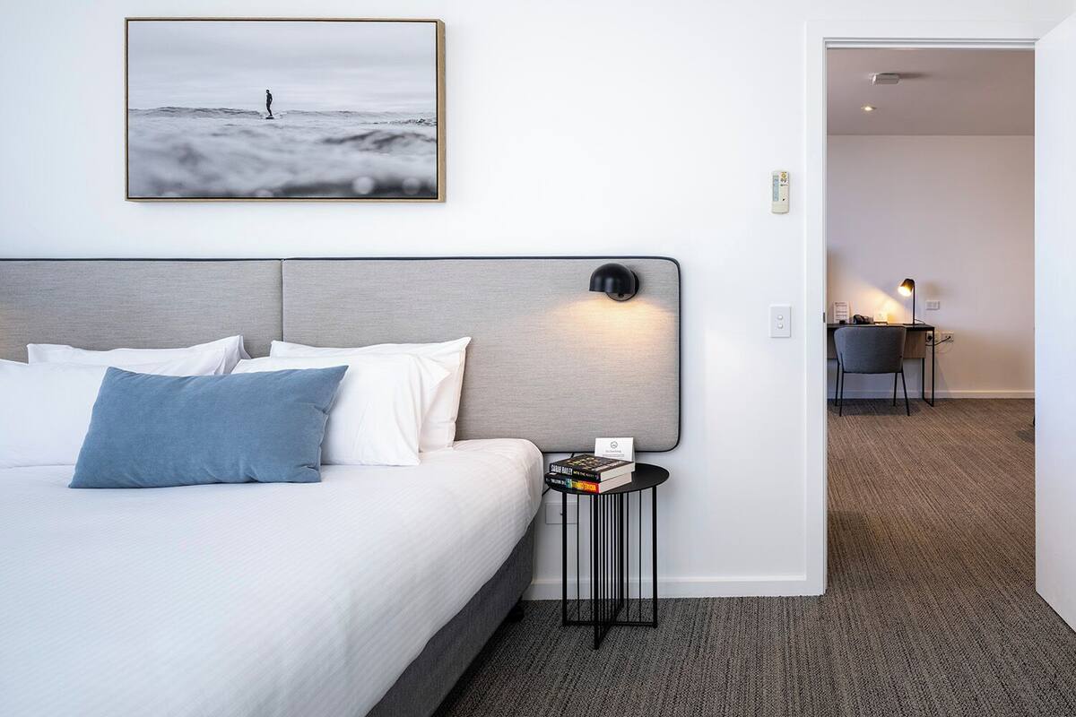 Habitación de hotel con cama, mesita de noche con lámpara, un cuadro en la pared y un escritorio con silla al fondo.