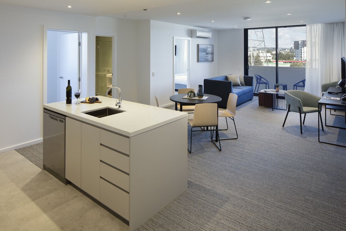 Una cocina moderna con una isla central, un comedor con una mesa redonda y sillas, una sala de estar con sofá y sillas, y un ventanal con vistas a la ciudad.