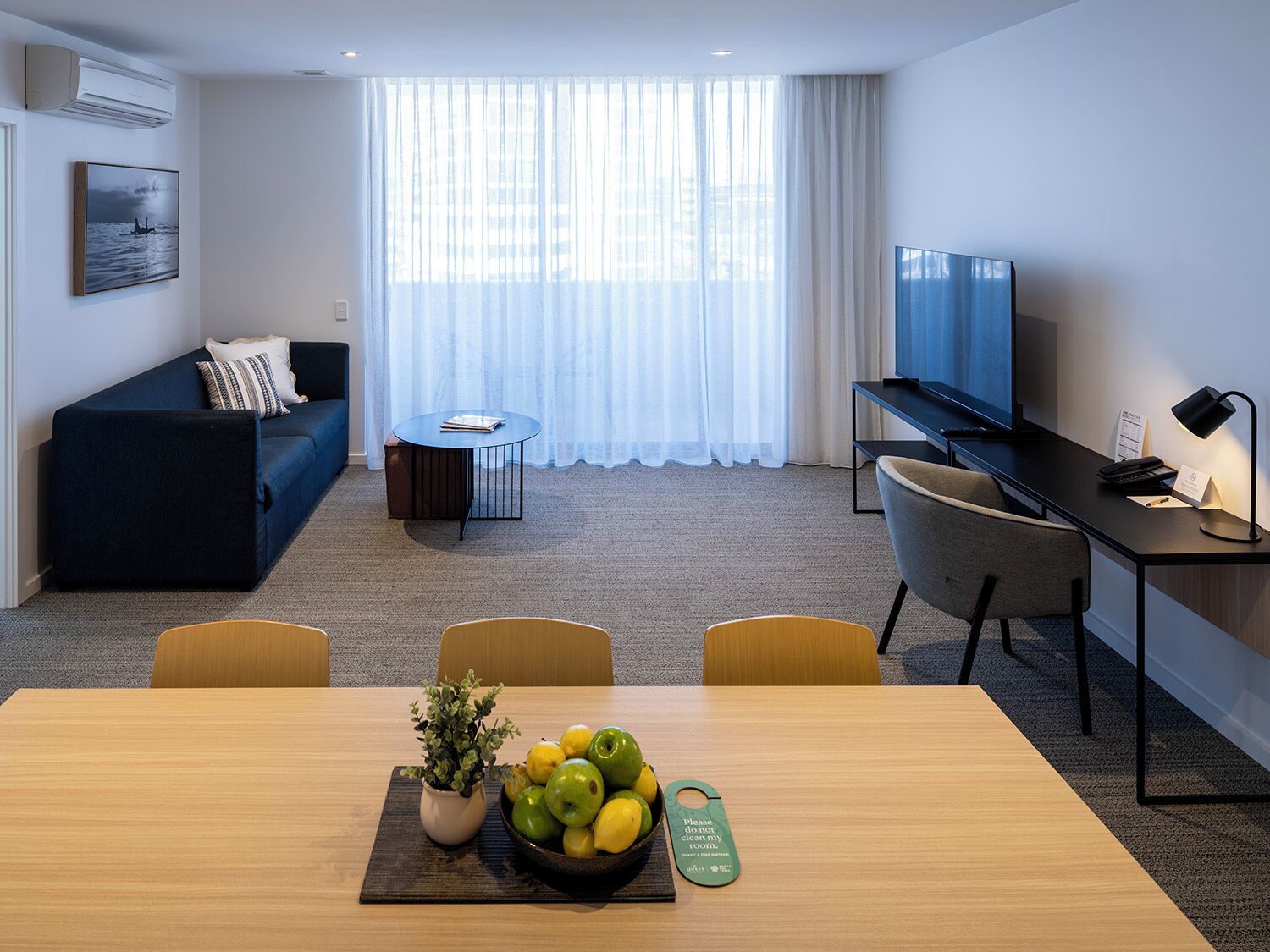 A modern hotel room with a sofa, a desk with a TV, a dining table with fruit and a plant, and a large window with sheer curtains.