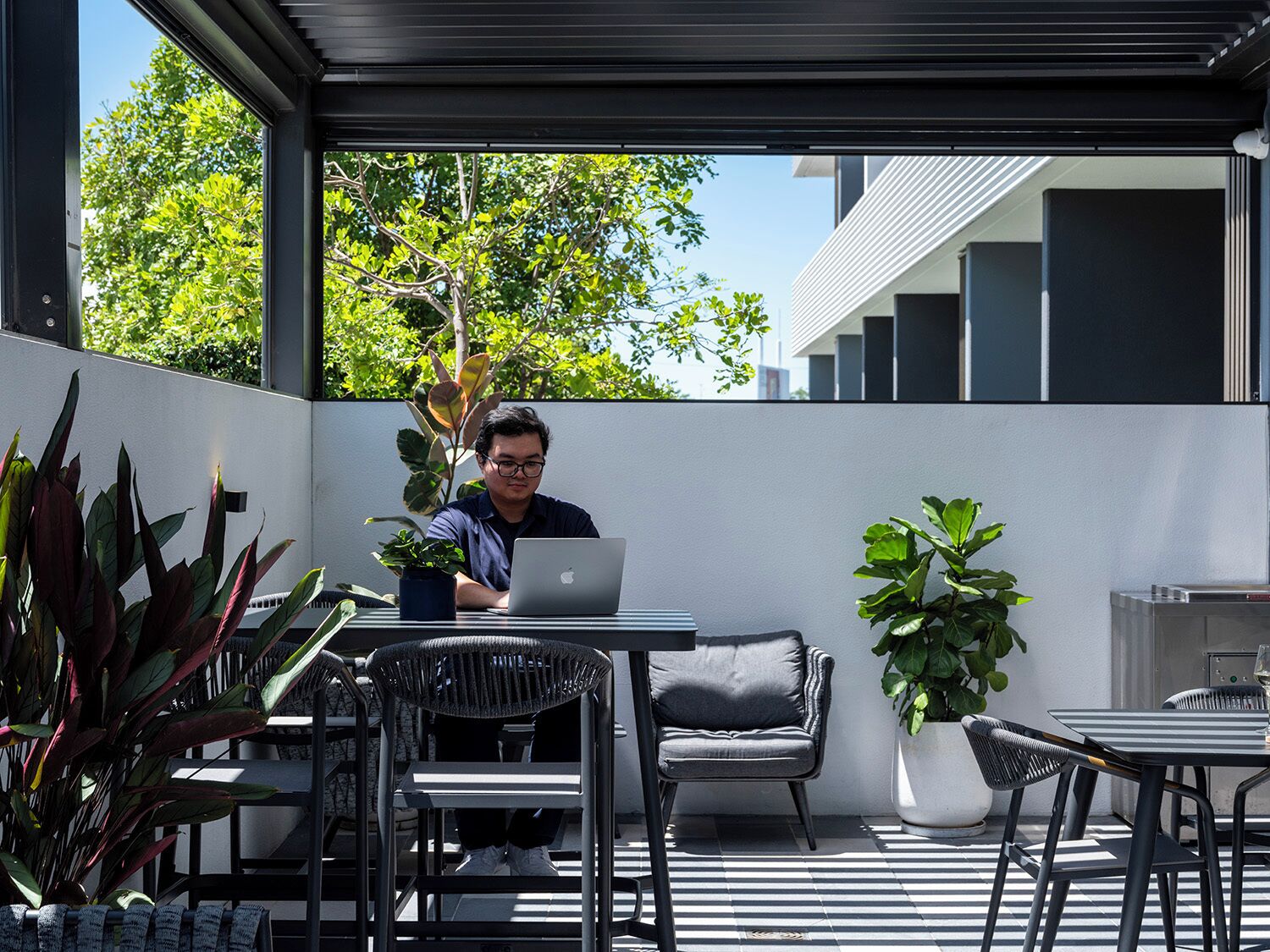 A man working on a laptop in an outdoor patio area with modern furniture and potted plants.