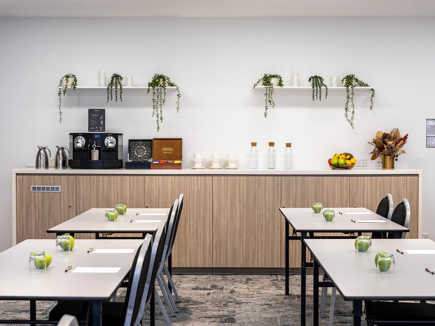 A modern office space with two long tables, chairs, a coffee machine, a tea set, and decorative plants.