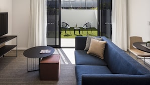 A modern living room with a blue sofa, a coffee table, and a TV on a stand. There is a sliding glass door leading to a balcony with outdoor seating. - Quest Robina (Robina)