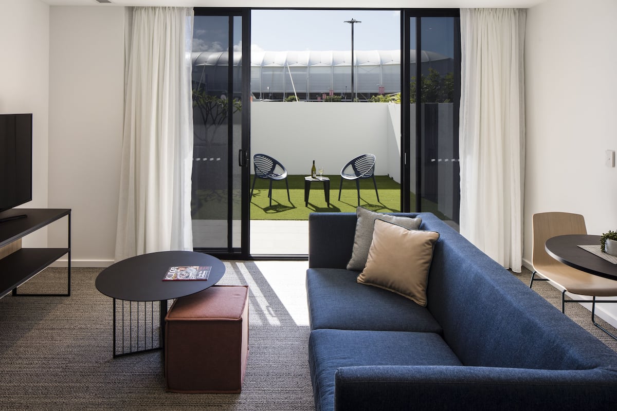 Una sala moderna con un sofá azul, una mesa de centro y un televisor sobre un mueble bajo. Hay un ventanal corredizo que da a un balcón con mobiliario exterior.