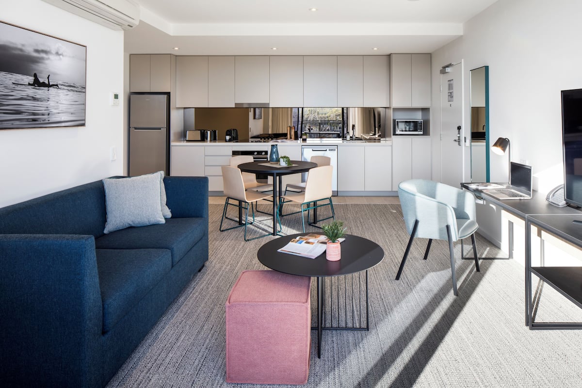 Una sala de estar moderna con sofá, reposapiés y mesa de centro, un comedor con sillas y una cocina con armarios y electrodomésticos.