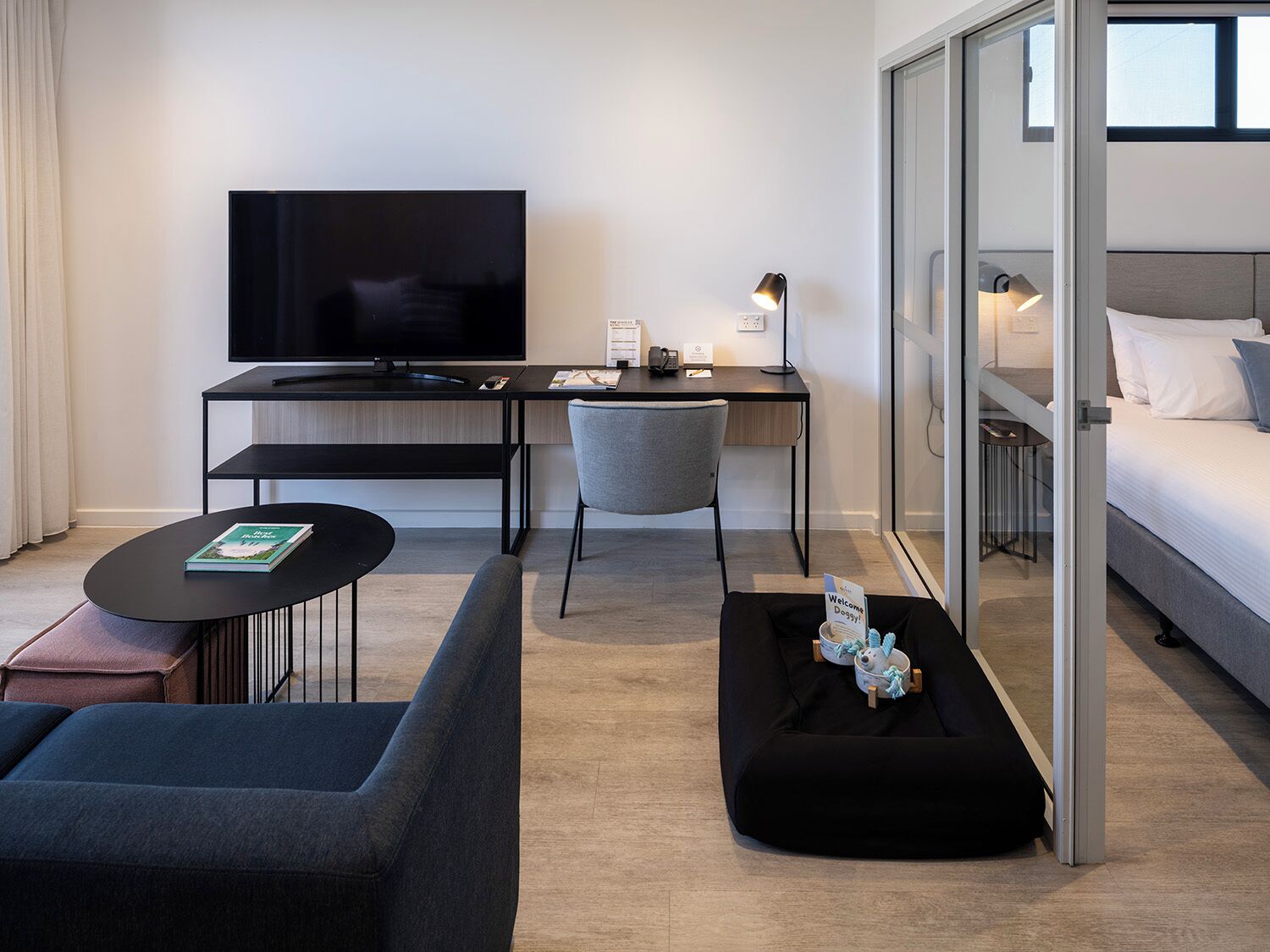 A modern hotel room with a flat-screen TV, a desk with a lamp, a sofa, a small table with a book, and a bed with a pillow.