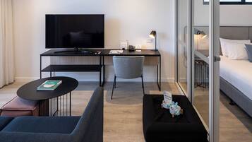 A modern hotel room with a flat-screen TV, a desk with a lamp, a sofa, a small table with a book, and a bed with a pillow.