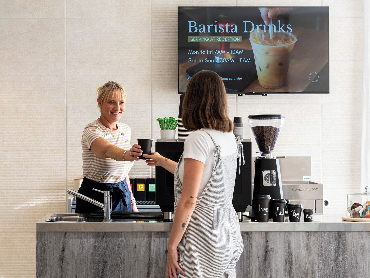 Eine Barista bedient einen Kunden am Verkaufstresen eines Cafés, über dem eine Schautafel mit dem Namen 'Barista Drinks' und den Öffnungszeiten angebracht ist.