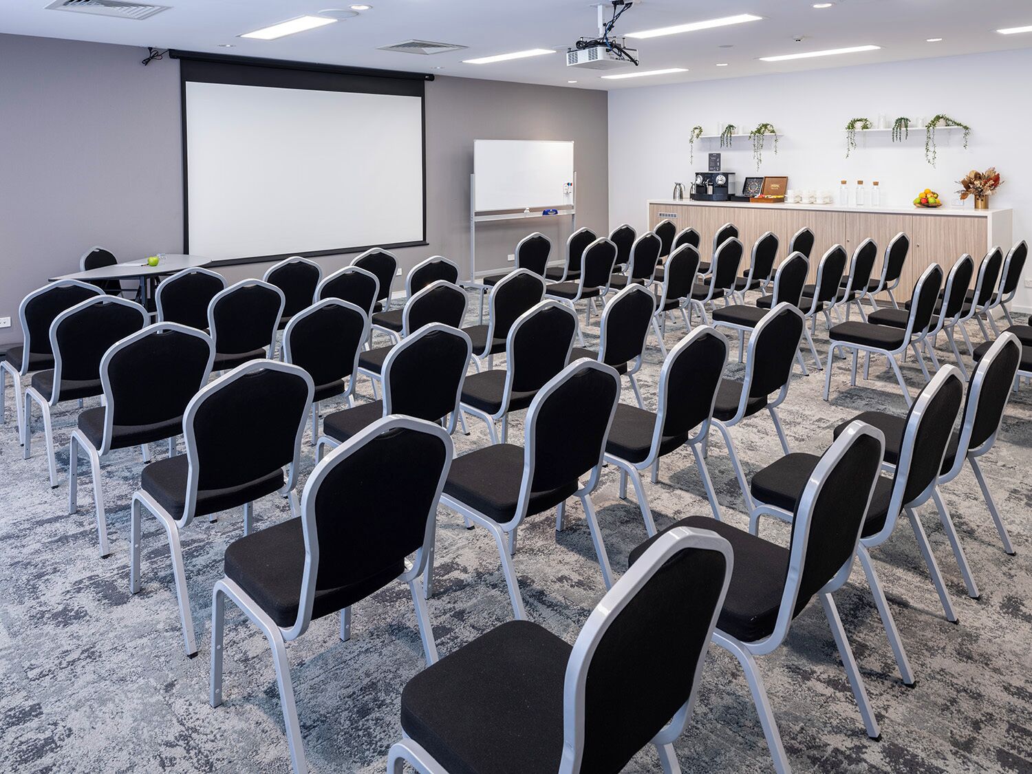 Une salle de conférence avec des rangées de chaises noires, un écran de projection, un tableau blanc et un comptoir d’accueil agrémenté de plantes et d’éléments décoratifs.