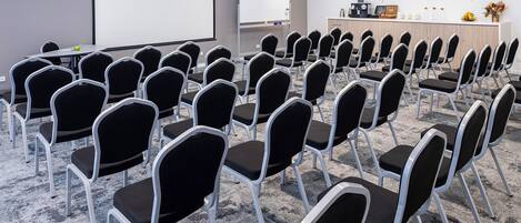 Une salle de conférence avec des rangées de chaises noires, un écran de projection, un tableau blanc et un comptoir d’accueil agrémenté de plantes et d’éléments décoratifs.