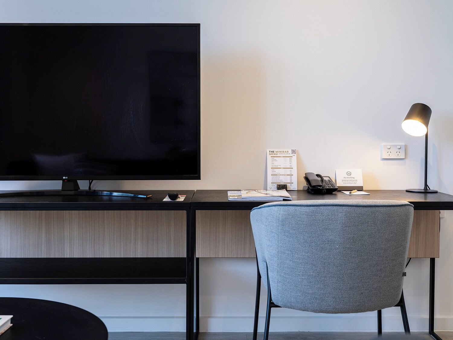 A modern hotel room with a flat-screen TV, a desk with a lamp, a chair, and a minibar sign.