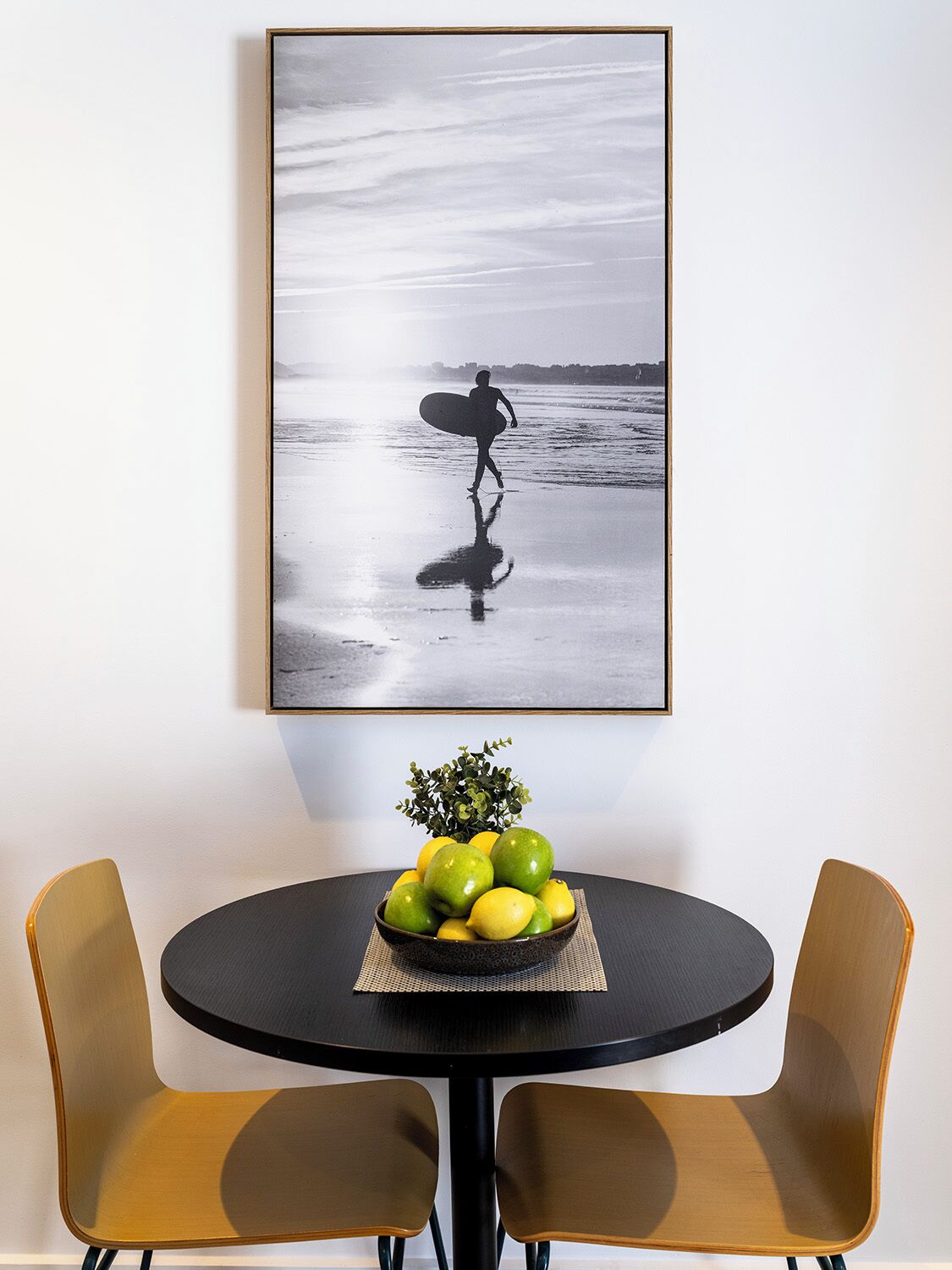 Ein gerahmtes Strandbild mit einer Person, die einen Surfboard trägt, einem Tisch mit einer Obstschale und zwei Stühlen.