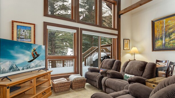 A cozy living room with a large TV, comfortable seating, and a view of a snowy deck. - Steps from Shuttle, Garage, 4 Bedroom + Basement, Private Scenic Setting (Vail)