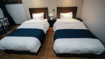 A hotel room with two beds, each with white bedding and a dark blue accent. There is a nightstand between the beds with a lamp and a phone.