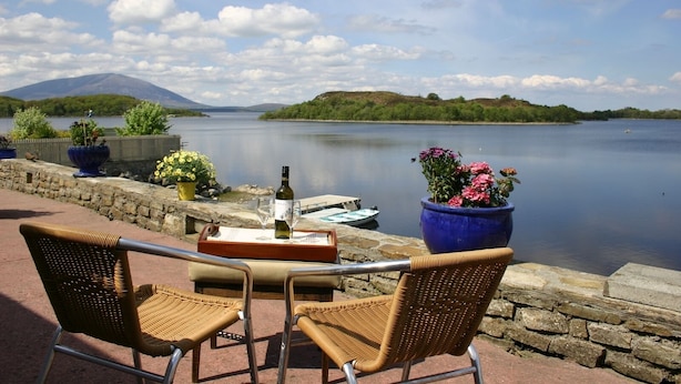 Een terras aan het meer met rieten stoelen, een tafel met fles en glas, en bloempotten.