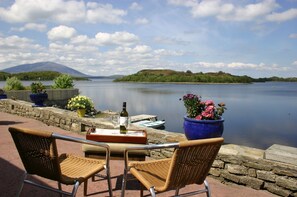 Un patio junto al lago con sillas de mimbre, una mesa con una botella y un vaso, y macetas con flores.