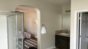 Una stanza con porta doccia in vetro, un letto con copriletto decorato, un asciugamano bianco appeso a un supporto, un lavandino con specchio e un tappeto decorativo sul pavimento.
