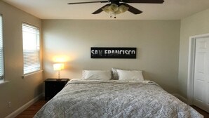 Una camera da letto con un letto, un comodino, una ventola a soffitto, un quadro appeso al muro e una finestra con persiane.