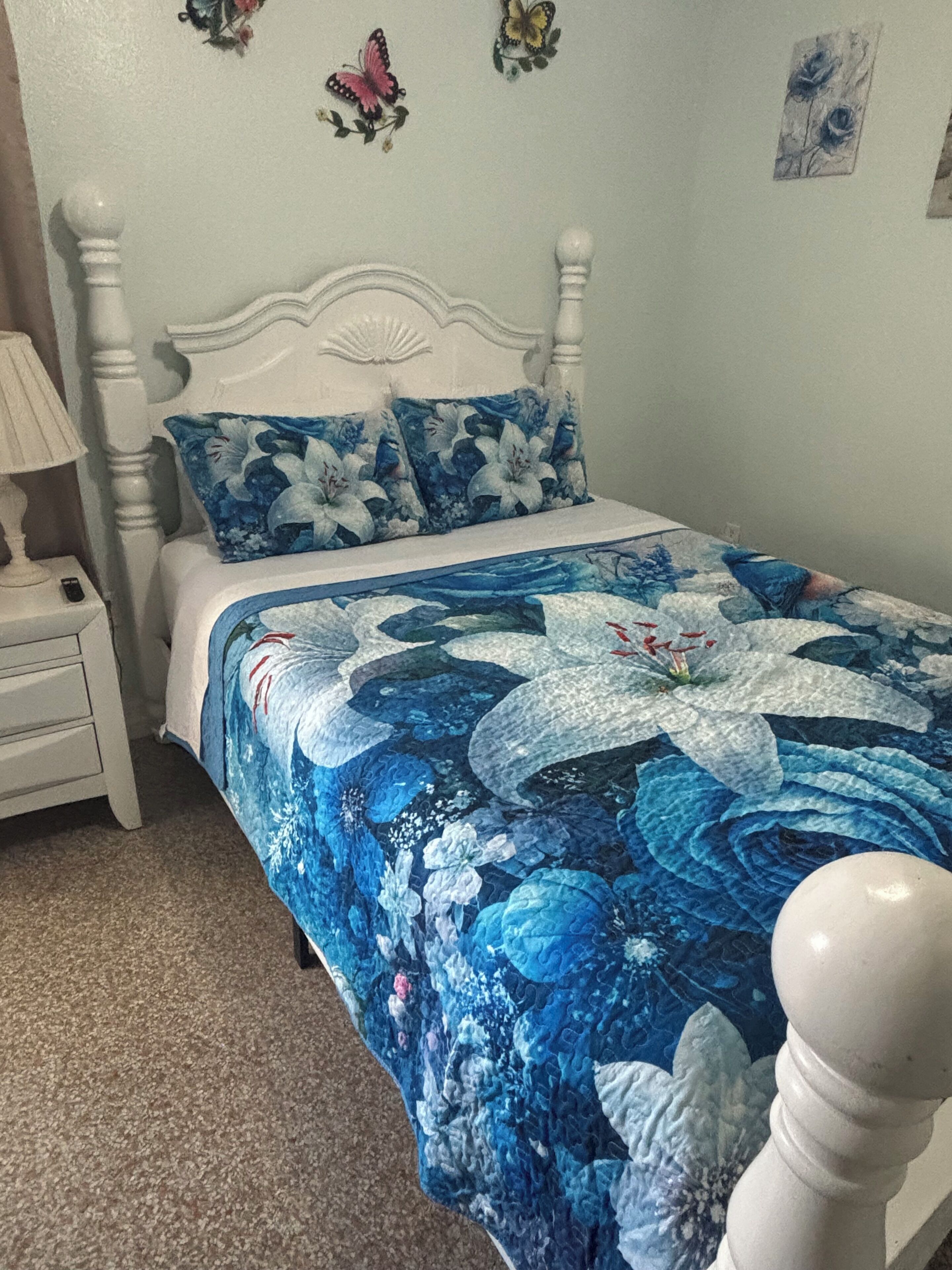 Uma cama com colcha florida, cabeceira branca e mesa de cabeceira com abajur.