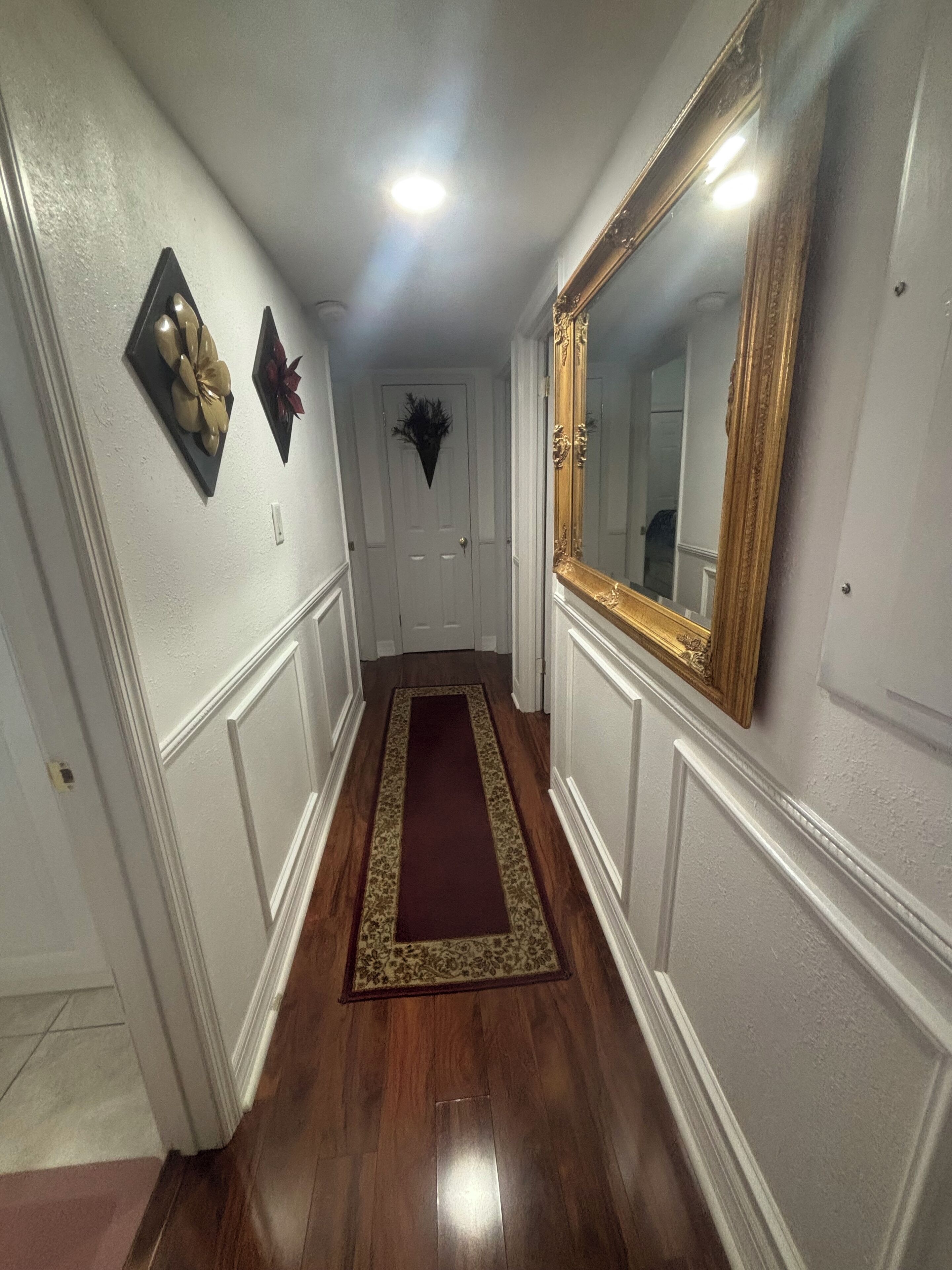 A hallway with a wooden floor, white walls, a dark red carpet, and a large mirror with a gold frame.