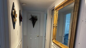 Um corredor com piso de madeira, tapete vermelho, paredes brancas e um espelho grande com moldura dourada.