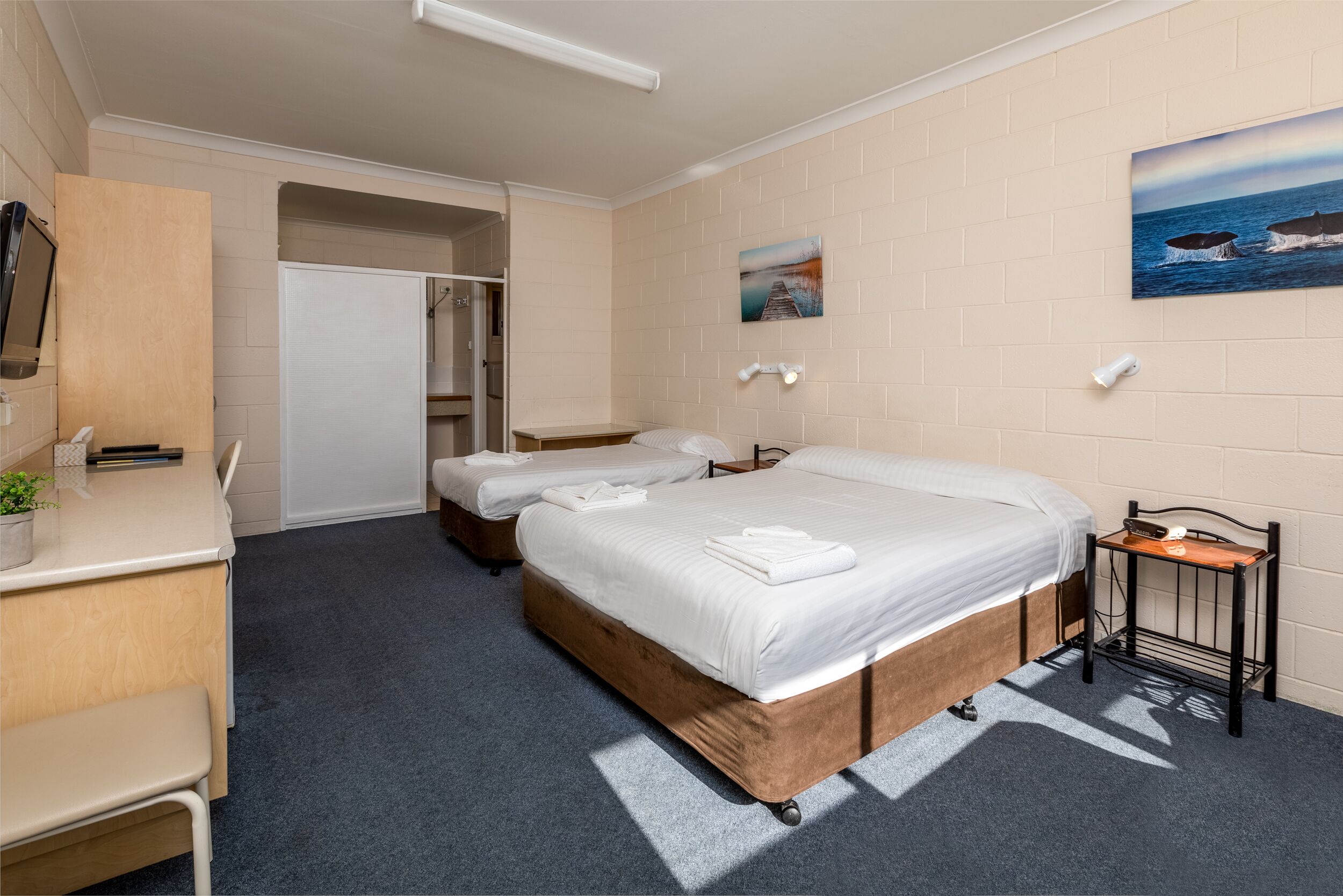 A hotel room with a large bed, two smaller beds, a desk, a chair, a wardrobe, and a painting of whales on the wall.