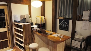 Un mostrador de recepción con una computadora portátil, documentos y un teléfono. Hay dos taburetes y un estante de madera con zapatos. Un calendario y un cartel que indica zona de empleados están en la pared.