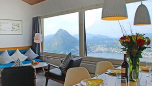Un comedor con una mesa dispuesta para dos personas, un sofá con cojines y vistas a una montaña y un pueblo a través de amplios ventanales.
