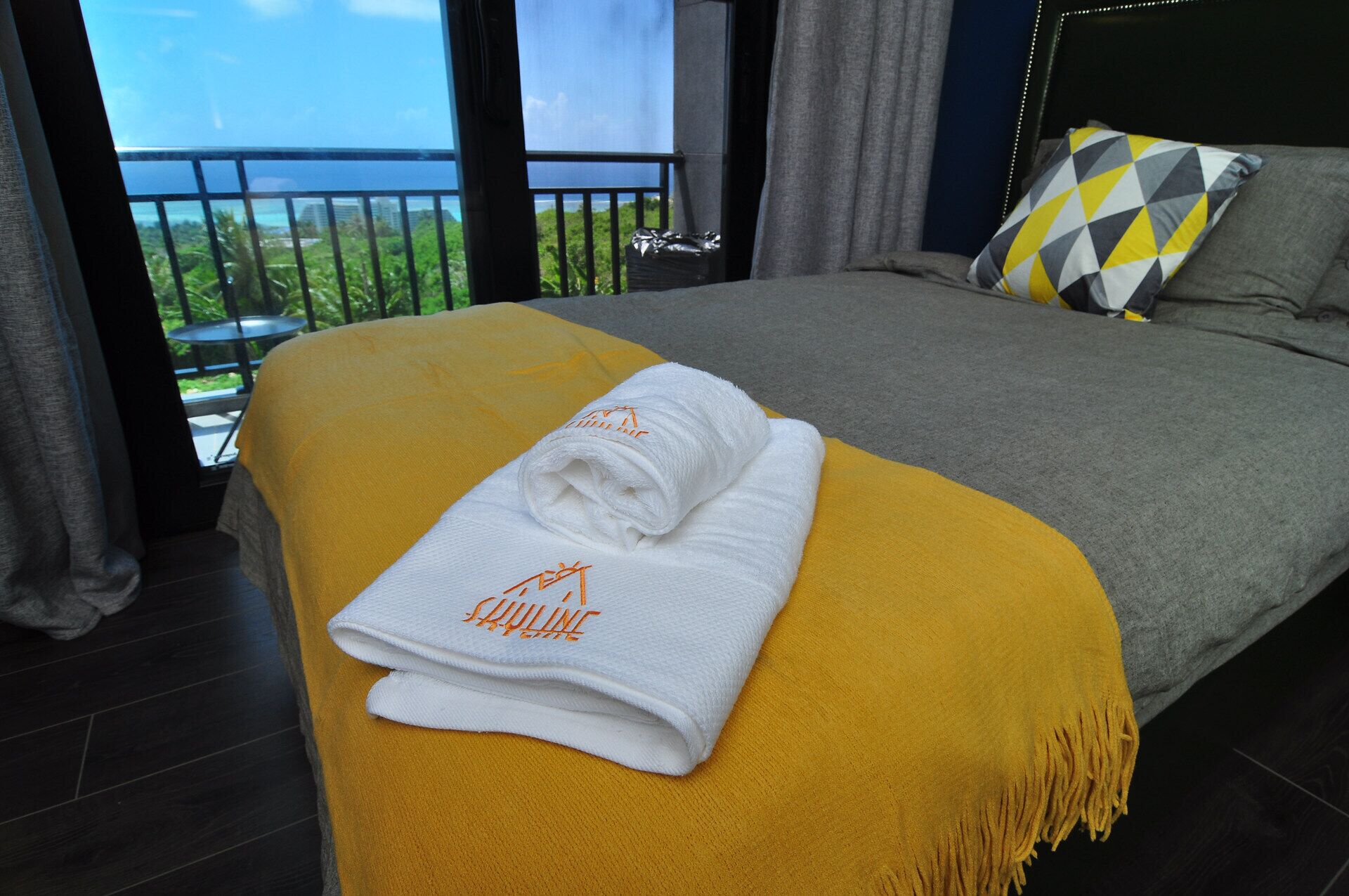 A neatly made bed with a yellow throw blanket and a folded white towel with a logo on a dark wooden floor.