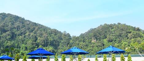 A poolside area with lounge chairs and blue umbrellas, surrounded by lush greenery and hills.