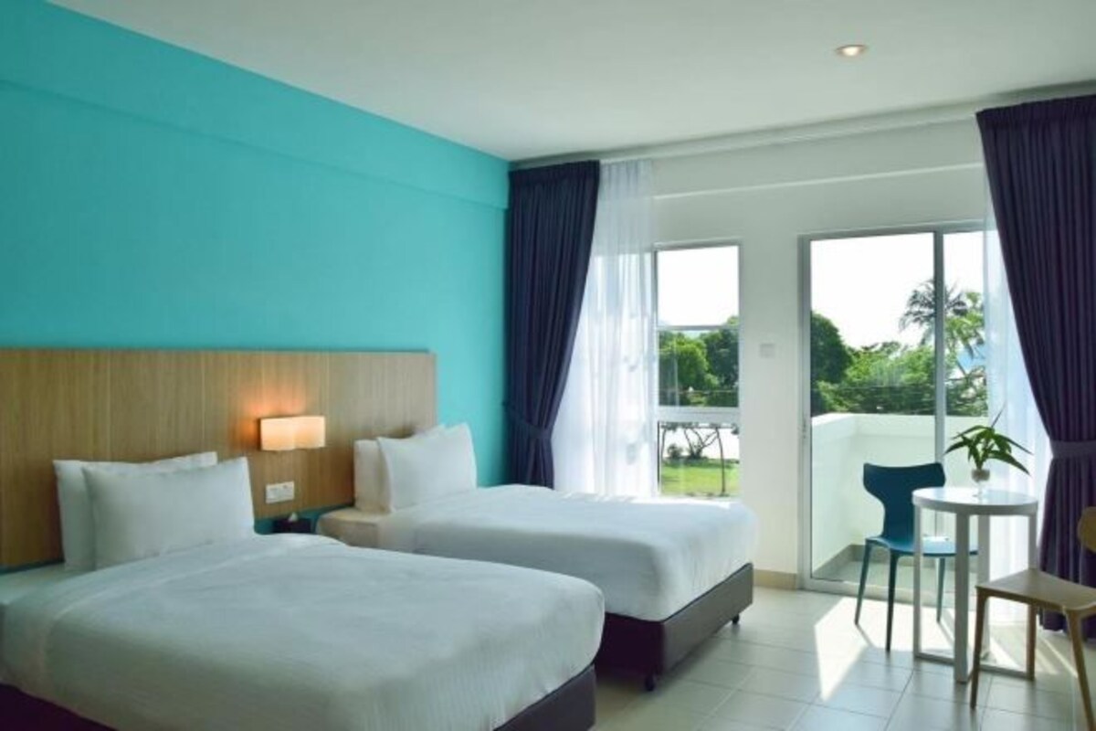 A hotel room with two beds, a small table, a chair, and a balcony with a view of greenery.