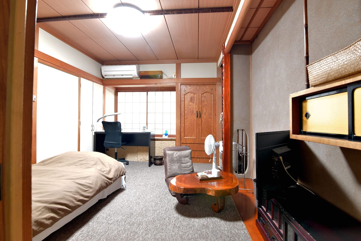 Habitación de hotel pequeña con una cama, un escritorio con silla, una mesita con ventilador y un televisor.