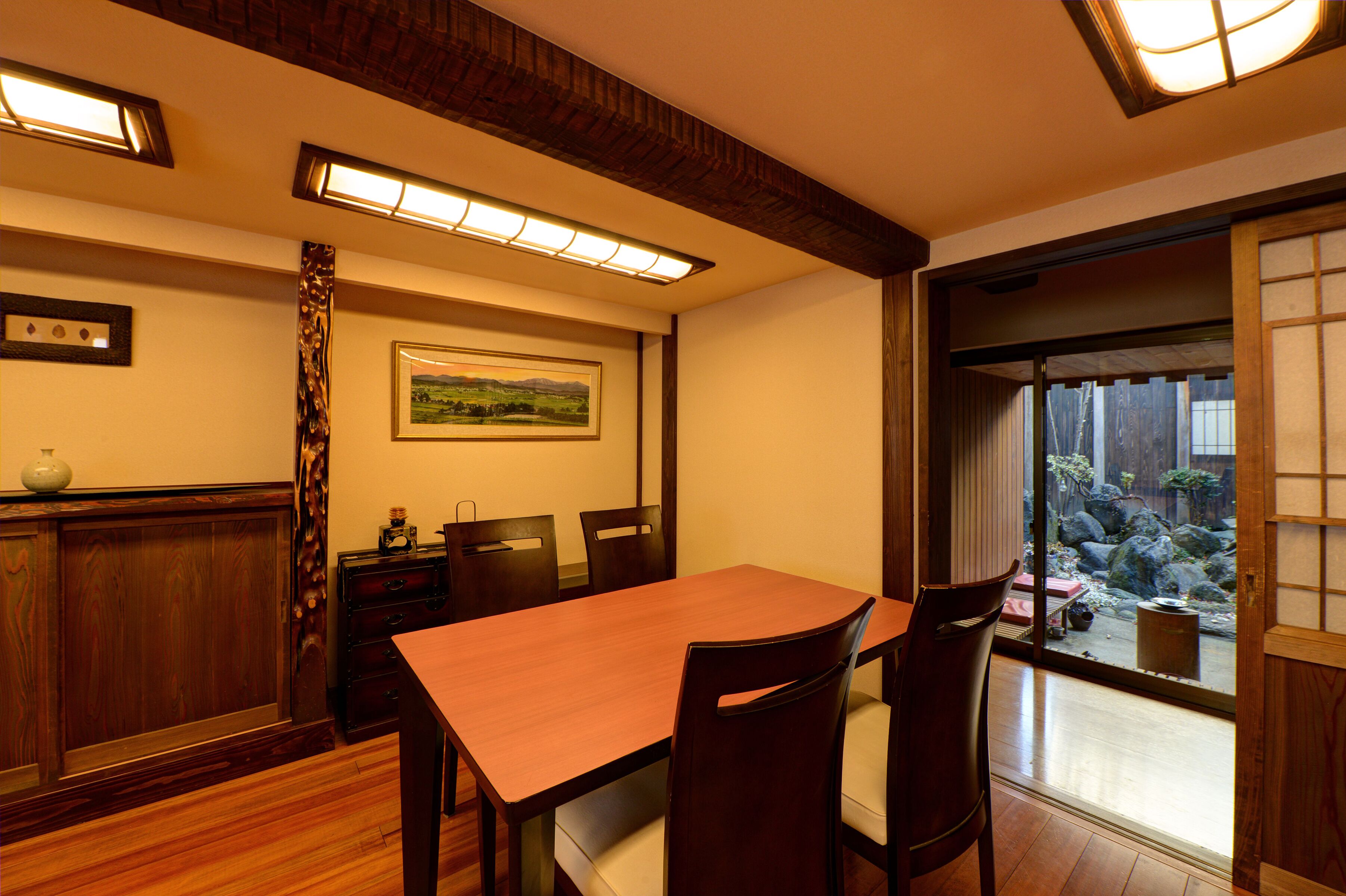 A dining area with a wooden table and chairs, a decorative vase, and a framed landscape painting on the wall.