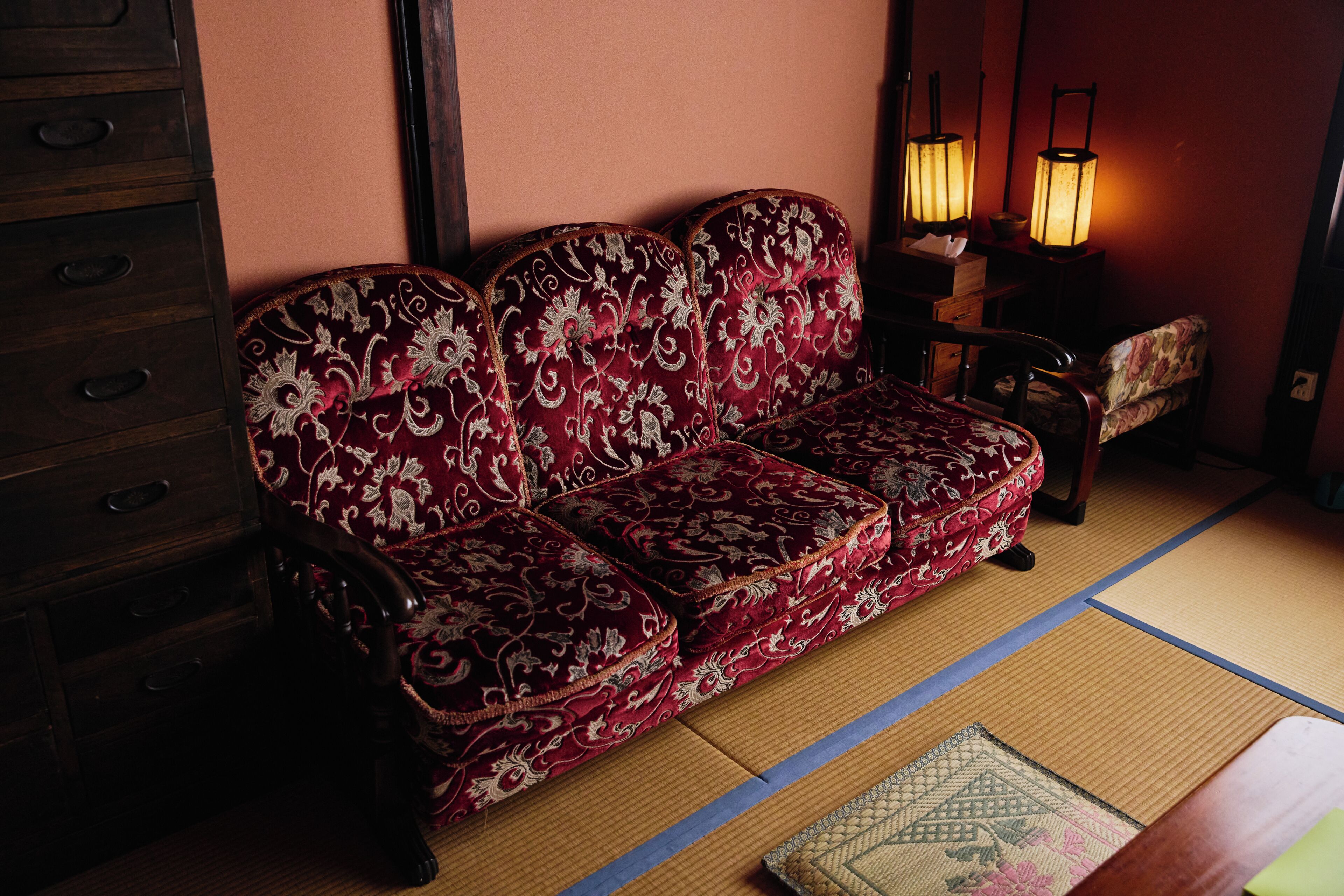 A traditional Japanese room with tatami flooring, a red patterned sofa, a wooden cabinet, and two floor lamps.