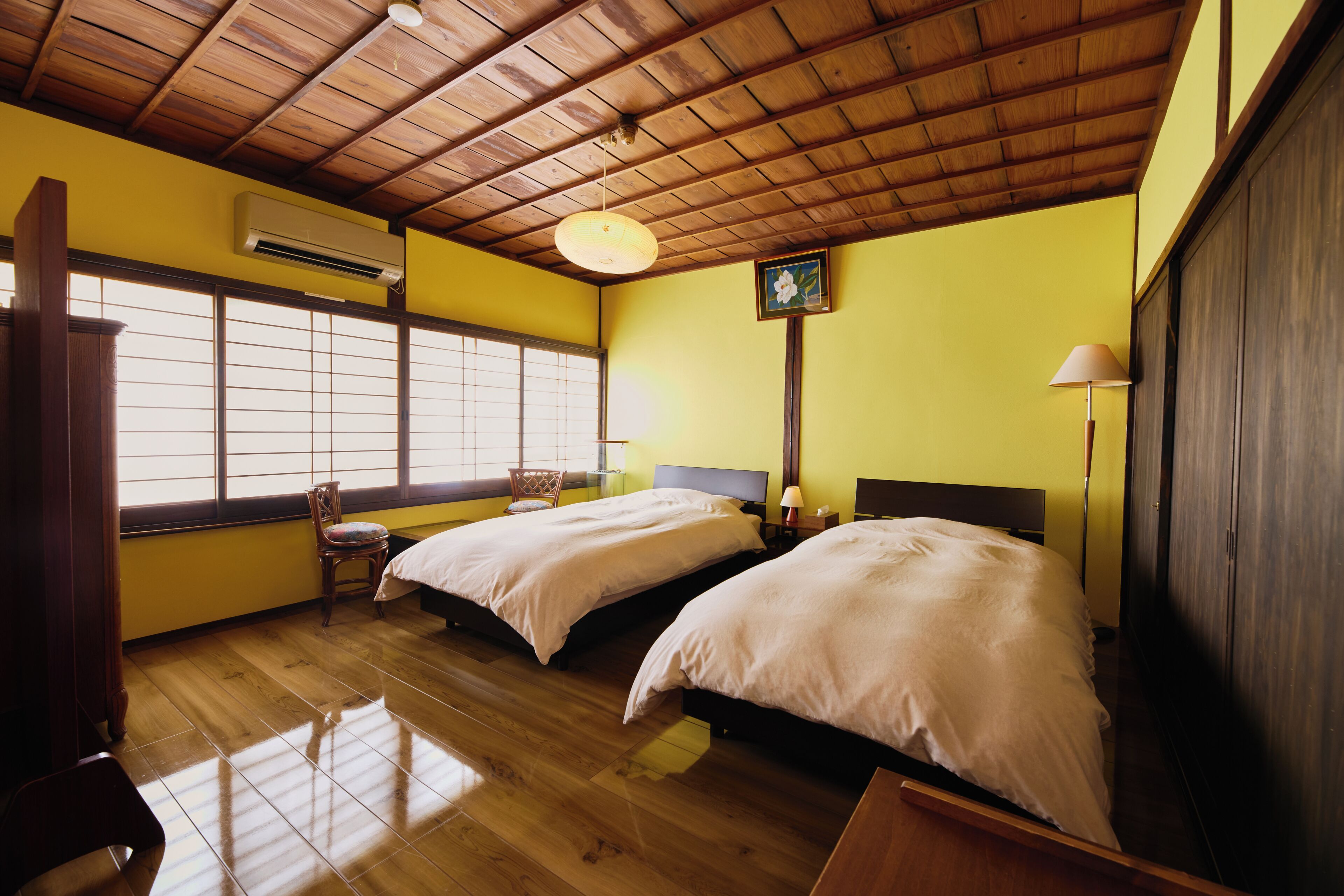 A room with wooden flooring, two beds, a chair, a window with blinds, an air conditioning unit, and a lamp.